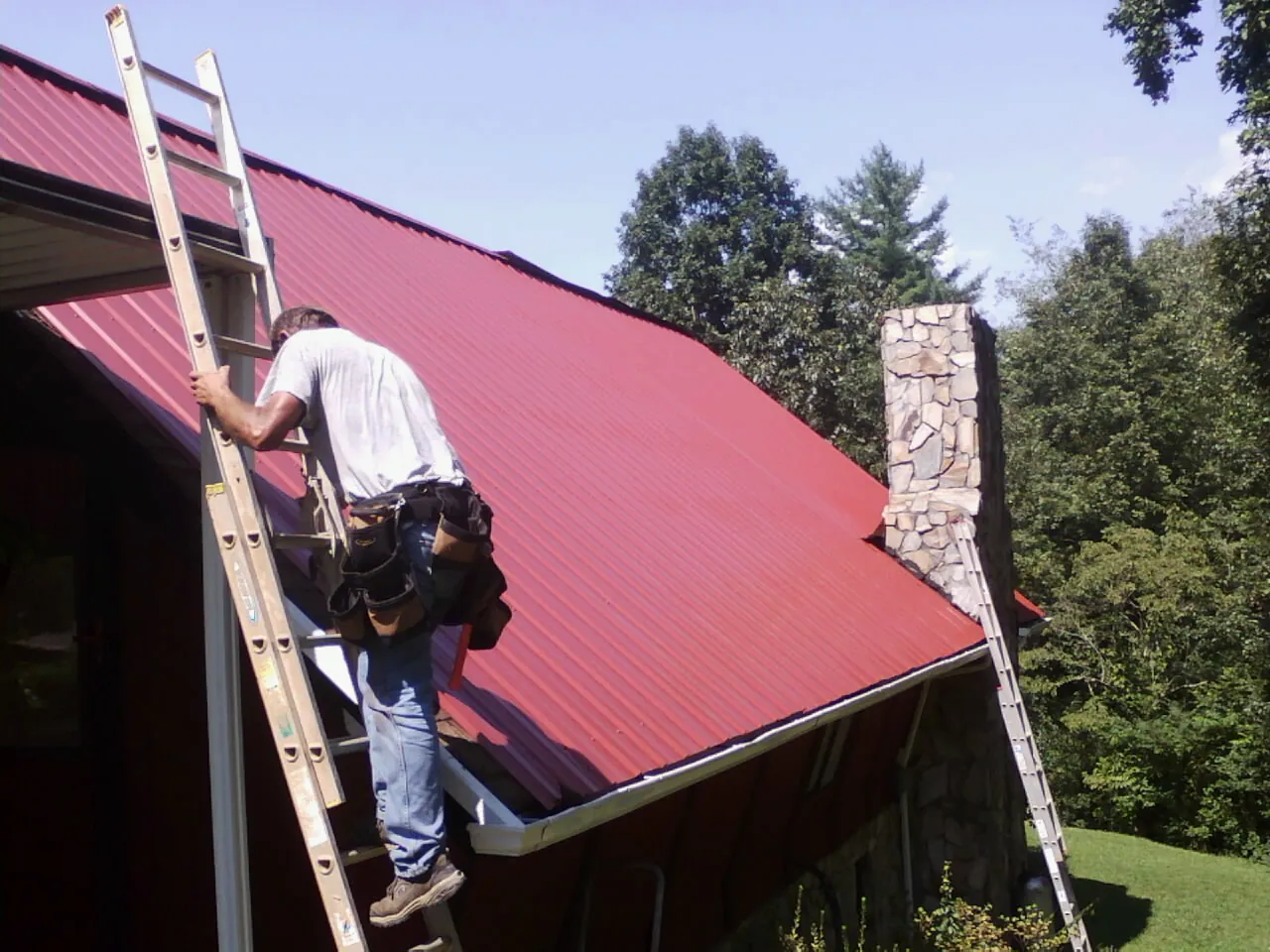 Roofer on a ladder beside a red metal roof and stone chimney — Soffit & Fascia Repair by Roofing Brothers in Haysville